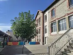 On voit au premier plan, derrière des barrières, une cour d'école avec un petit arbre et un muret coloré un peu plus loin. À gauche on devine un préau. Au second plan se dresse le bâtiment qui abrite l'école en elle-même ainsi que la mairie du village au dernier étage. L'ensemble est plutôt gris mais de larges fenêtres font en sorte que le tout ne semble pas trop triste.