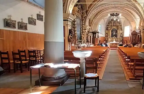Photographie de l'intérieur d'une église avec les colonnes, les autels et les bancs ; on voit un maître-retable au fond de l'église ainsi que les différents ornements de l'édifice qui semblent baroques et gothiques au vu de leurs décorations opulentes pendant qu'une messe se déroule dans l'église en présence de fidèles.