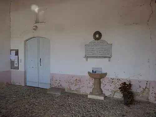 Le porche avec de gauche à droite la porte, la pierre des « excommuniés » ou des « cagots », un monument aux morts et des fonts baptismaux intégrés dans le mur.