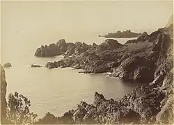 Photographie noir et blanc. Littoral avec au second plan la mer et une île.