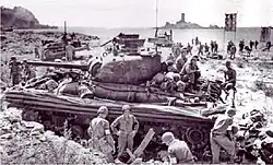 Photo noir et blanc. Au premier plan un char amphibie avec sa jupe sur une plage en appui des troupes débarquant. Au loin une île.