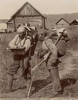 Photographie en noir et blanc de deux hommes avec des fusils devant des habitations.