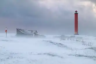 Phare rouge se dressant à droite au second plan dans un paysage enneigé, balayé par le fond, avec des nuages gris. Des bâtiments d'un étage en bois émergent au centre de la brume.