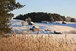 Photographie au premier plan d'un lac gelé avec un esker le bordant sur lequel se trouvent quelques maisons en bois, le tout dans un passage hivernal.