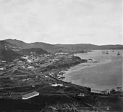 Photographie en noir et blanc d'une petite ville installée sur des monts au bord d'une baie où se trouve des bateaux.