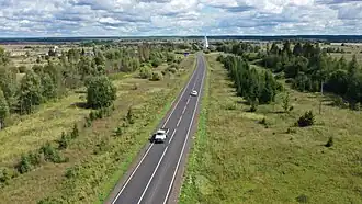 Photographie d'une route traversant un paysage mixte de champs et bosquets, avec un village possédant une église en arrière-plan.