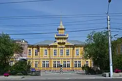 Bâtiment postal russe datant de l'Empire russe de deux étages, caractéritisque par sa peinture en jaune vif. Il est situé sur une place et arbore un toit pointu caractéristique. Des fenêtres rectangulaires, nombreuses et régulières, illuminent la façade. Un panneau bleu indique "Pochta Rossii", le service postal russe. Des arbres verdoyants bordent la place et le tout est sur fond d'un ciel bleu clair.