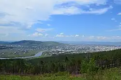 Vue depuis une colline verdoyante d'une plaine urbanisée dans laquelle coule une rivière. Au fond se distingue une colline avec le ciel bleu et des nuages.