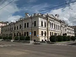 Bâtiment de deux étages, de couleur jaune clair, situé à l'angle de deux rues et occupe une position dominante dans le paysage urbain. De style classique, avec des fenêtres rectangulaires, un toit plat et des éléments décoratifs simples, il est entouré d'arbres et de buissons. Le ciel est bleu et quelques nuages sont visibles.