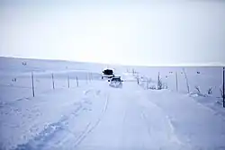 Une route au centre, avec moins de neige que le reste de l'image, file vers l'arrière-plan, dans un paysage enneigé valloné. Deux voitures se croisent au centre de l'image sur la route, chacune dans un sens.
