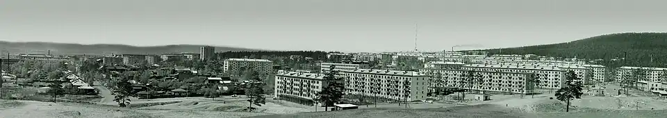 Photographie en noir et blanc, vue aérienne d'une ville en pleine expansion. Au premier plan l'on distingue des immeubles résidentiels de style soviétique (khrouchtchevka), alignés de manière ordonnée. En arrière-plan, une forêt dense borde la ville, tandis qu'un ciel nuageux domine la scène.