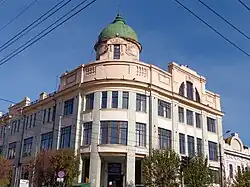 Photographie en contre-plongée montrant un bâtiment aux façades, de couleur pastel, rythmées par de nombreuses fenêtres. Un dôme vert coiffe le bâtiment, sur fond de ciel dégagé.