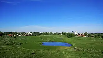 Vue d'une prairie avec un petit étang et des églises et maisons en arrière-plan.