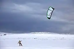 Une personne glisse sur la neige en bas à gauche de l'image, tirée par un grand cerf-volant blanc en haut à droite, dans un paysage blanc et venteux, avec des nuages gris en arrière-plan.