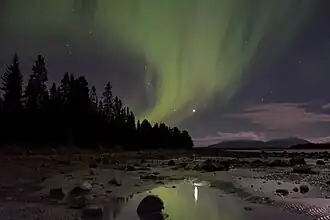 Vue depuis une plage de graviers lors de la nuit polaire d'une aurore boréale verte en centre-haut de l'image. Au centre-gauche, une forêt de conifère se trouve, ainsi que des monts au loin au centre-droit. L'aurore boréale se reflète dans les flaques d'eau de la plage en bas au centre.