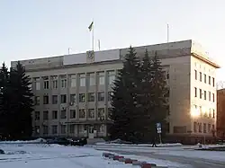 Photographie en couleurs. Bâtiment en pavé droit haut de trois et surmonté d’un drapeau ukrainien. Des conifères masquent partiellement la façade et de la neige recouvre le sol. Ciel dégagé.