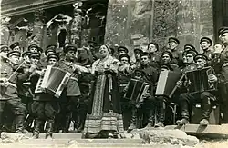 Photographie en noir et blanc d'une chanteuse en costume traditionnel accompagnée d'un orchestre militaire et chantant devant des ruines.