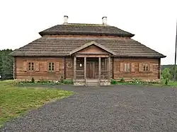 Photographie d'une maison en bois avec un toit en chaume. Un porche et quatre fenêtres se trouvent sur la façade avant