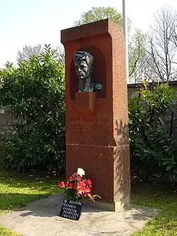Photographie d'un stèle funéraire rectangulaire en tuf orange au centre de la laquelle repose un buste de Missak Manouchian.