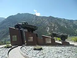 Photographie du monument de la légende des années 1930. Quatre blocs portent en tout deux véhicules, avec les blocs qui montent en hauteur. Le premier véhicule est plus imposant que le second.