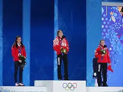Trois biathlètes sur le podium, chacune tenant un bouquet de fleur dans les mains.