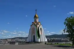 Photographie d'un bâtiment en forme de cône sur une place, surmontée d'une coupole dorée avec une croix orthodoxe. Au fond se distingue une ville et des collines, sur fond de ciel bleu.