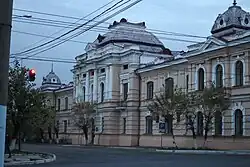 Photographie d'un bâtiment de style classique de deux étages avec un centre couvert d'une coupole verte, sous un ciel nuageux avec une rue vide.