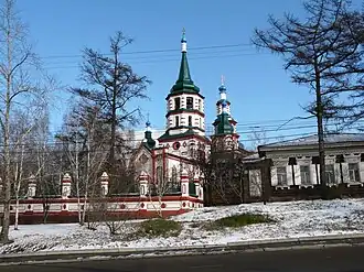 Église aux murs blancs soutenus de rouge et aux toits vert foncé, dans un paysage enneigé et arboré.