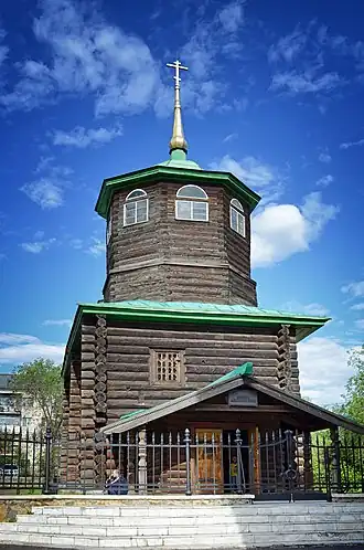 Photographie de face d'une église en rondins de bois, composée d'un premier étage en cube, sur lequel se trouve une tour en octogone surmontée d'une flèche dorée avec une croix orthodoxe.