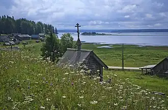 Vue depuis une petite colline d'une petite chapelle en bois en premier plan d'architecture russe avec une croix orthodoxe à son sommet. En second plan à gauche se distingue un village avec des maisons en bois, et en arrière-plan le lac avec au fond des collines recouvertes de taïga.