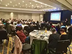 Photo de personnes assises à des tables regardant une présentation sur un écran.