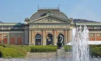 Photo de l'entrée d'un bâtiment : l'entrée est composée de trois grandes portes identiques, séparées par des formes de colonnes carrées.