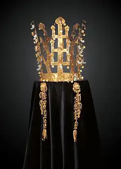 Couronne faite de bandeaux en or en forme d'arbres dressés aux feuilles d'or et pendentifs de jade (en forme de virgule). Photographie de face, sur fond sombre, la couronne étant posée sur un velours noir.