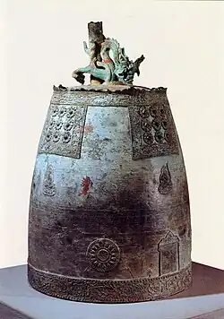 Photographie d'une grande cloche en métal. Un dragon est visible à son sommet, et sa surface comprend plusieurs motifs décoratifs. Elle est siuée sur un socle dans un musée.