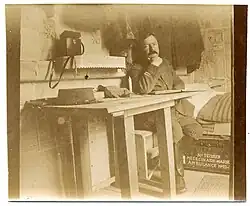  photo de couleur sépia montrant un médecin en uniforme avec galons de lieutenant assis dans un abri servant de poste de secours, sous son lit on voit sa cantine peinte à son nom et son grade