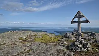 Une croix en bois qui ressemble à une croix orthodoxe depuis le sommet d'une montagne dénudée de végétation, avec un panorama grand avec un lac et des étendues de taïga en second plan.