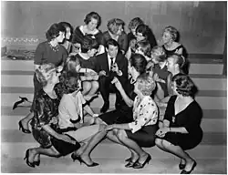 Photographie en noir et blanc représentant, sur des marches, Jacques Brel assis au centre, entouré d'une dizaine de jeunes femmes toutes tournées vers lui.