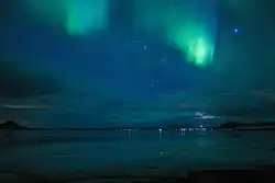 Aurore boréale verte dans un ciel bleu et noir. Au fond, l'île illuminée.