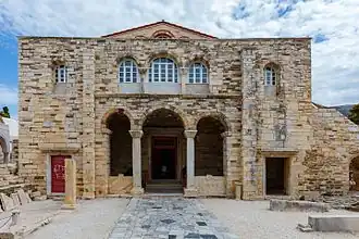 Photographie couleurs : un bâtiment religieux en pierres jaunes à toit de tuiles, précédé d'une cour