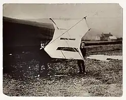 Photographie en noir et blanc de deux personnages portant un grand cerf-volant sur un terrain herbeux, légèrement accidenté.