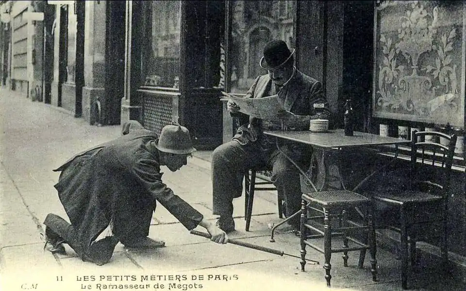 Un ramasseur de mégot exerçant devant la terasse d'un établissement à Paris, au tournant du XXe&nbsp;siècle.