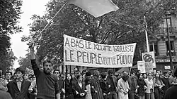 Photographie en noir et blanc montrant des personnes en train de manifester dans la rue. Des manifestants brandissent une banderole sur laquelle il est écrit « À bas le régime gaulliste, le peuple veut le pouvoir ».