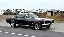 Voiture 3 portes noire aux angles marqués, la fenêtre latérale formant un trapèze presque régulier, l'avant long, le coffre plus court