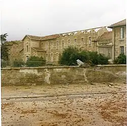 Photographie d'un grand bâtiment en pierre à la toiture partiellement détruite derrière un muret.