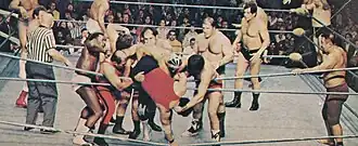 Photographie en couleurs d'un match de catch, où cinq catcheurs tentent de projeter un adversaire hors du ring.