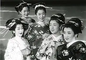 Cinq femmes souriantes prennent la pose, elles sont vêtues kimonos et leurs cheveux sont ornés de kanzashi (ornements traditionnels japonais) (image en noir et blanc).
