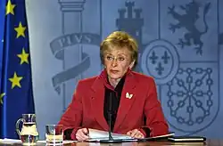 Photographie en couleur d'une femme vêtue d'une veste rouge. Elle est assise derrière un bureau. Devant elle est posé un microphone. Une cruche d'eau et un verre sont posés à sa droite. Sur sa droite est placé un drapeau bleu comportant des étoiles jaunes.