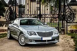 Une voiture grise 2 portes vue de l'avant droit devant une grille marquant l'entrée d'une propriété.