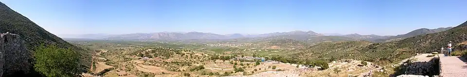 Panorama d'une plaine, prise depuis une hauteur. Aux extrémités gauche et droite, des flancs de collines. Au fond, après la plaine, une chaîne de montagnes basses. Au premier plan, les ruines de la cité de Mycènes.