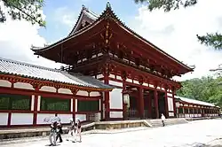 Porte dans le mur d’enceinte, aux poutres rouges et aux murs blancs. Toit à demi-croupes au faîte plat, courbé à l’extrémité.